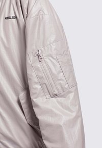 Svijetlosiva puffer jakna s sjajnom, naboranom teksturom. Ima zippered džep na rukavu i logo na gornjem dijelu ruke.
