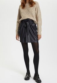 Svart läderkjol med knytbälte och fickor, kombinerad med en beige stickad tröja och svarta strumpbyxor, kompletterad med svarta loafers.