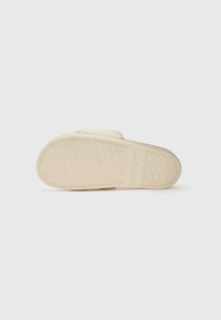 Light-colored sandal with a textured rubber sole, featuring a wide strap and embossed logo. The sole has a diamond pattern for grip.