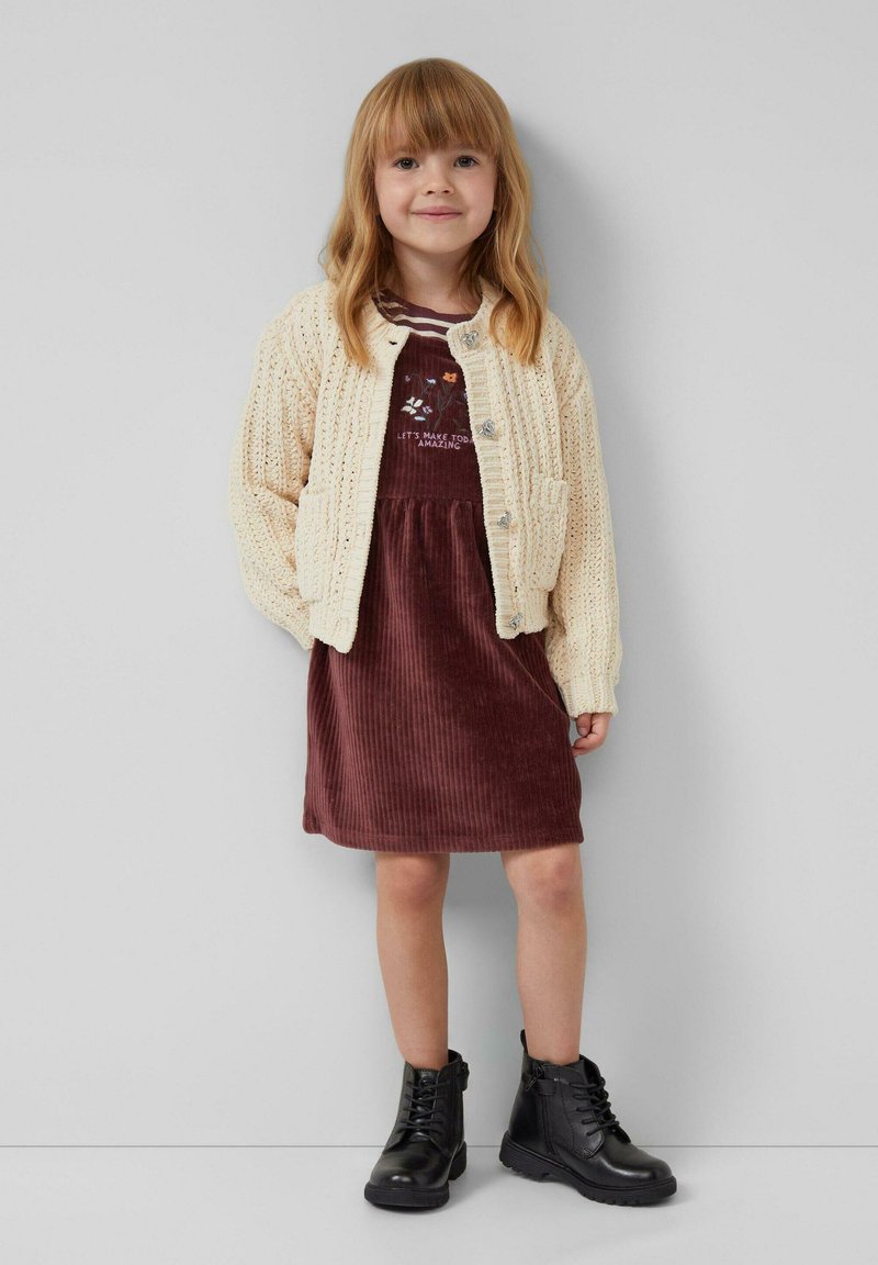 Cremiger Strickcardigan, kastanienbraunes Cordkleid mit geripptem Muster und schwarze Schnürstiefeletten. Einfaches Design mit Knopfdetails.
