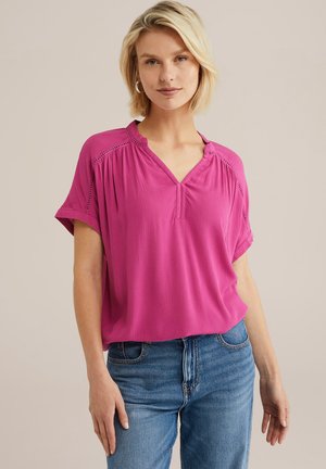Lyserød kortærmet bluse med V-hals og tekstureret stof. Har dekorativ syning langs skuldrene, parret med blå jeans.