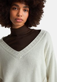 Strickpullover mit V-Ausschnitt in hellem Cremeweiß mit gerippten Details. Über einem dunkelbraunen Rollkragenpullover getragen, was die Textur und das geschichtete Design hervorhebt.