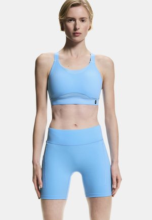 Junge Person trägt ein passendes hellblaues Sport-BH und hoch taillierte Radlerhose, steht aufrecht vor einfarbigem Hintergrund.