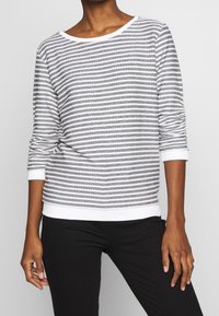 Haut à manches longues rayé gris et blanc avec encolure bateau et poignets blancs, fabriqué dans un tissu texturé avec une coupe décontractée.