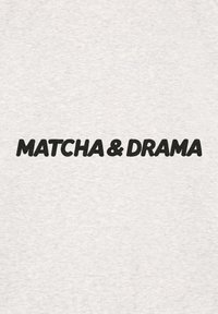 T-shirt grigio in cotone con testo nero in grassetto che recita "MATCHA & DRAMA." Il tessuto ha una consistenza liscia con un delicato motivo melangiato.