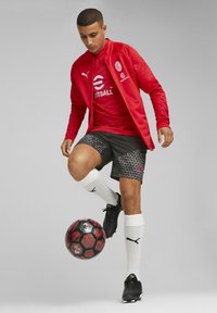 Rode jas met logo, gestreepte rode trui, zwarte shorts met geometrisch ontwerp, witte sokken, zwarte voetbalschoenen en een zwart-rode voetbal.