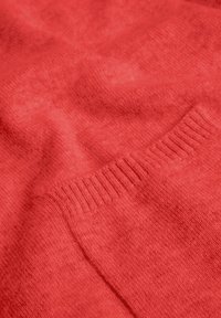 Pull en tricot corail, avec une texture douce, des poignets côtelés et un léger éclat. Le tissu présente un motif subtil pour un détail supplémentaire.