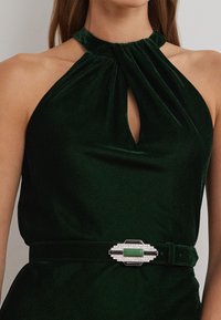 Zeleni žametni halter top s ključnim izrezom, opremljen srebrnim kopčanjem s zelenim kamenom in zapletenim detajlom.