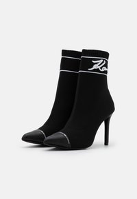 Botines de tacón alto negros con parte superior de punto, que presentan un logo blanco en el puño y punta afilada negra brillante. Tejido texturizado, tacón de aguja delgado.