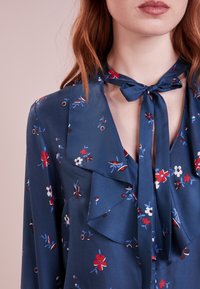 Granatowa bluzka w kwiatowe wzory w kolorach czerwonym, białym i niebieskim. Posiada wiązanie przy szyi oraz falbany z przodu. Gładka tekstura materiału.