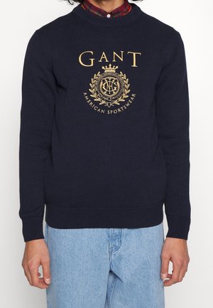 Mann trägt einen marineblauen GANT-Pullover mit goldenem Emblem und Text über einem rot-blau karierten Hemd, kombiniert mit hellblauen Jeans.