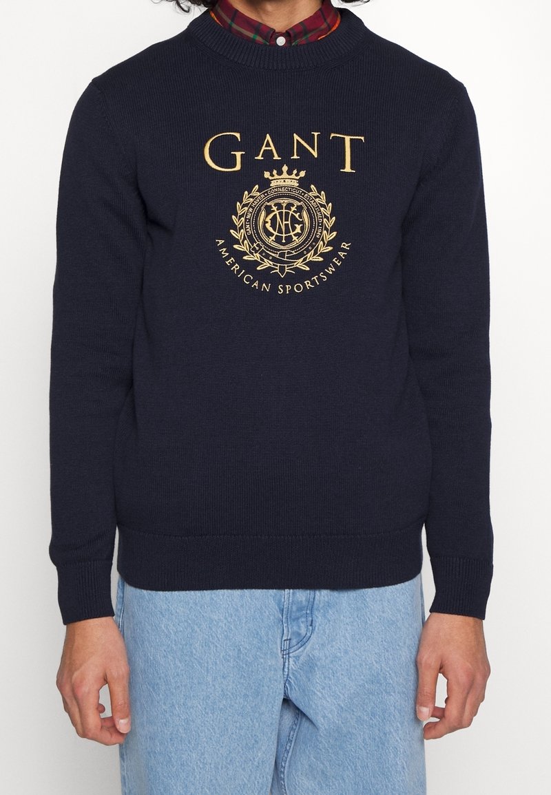 Mies käyttää tummansinisestä GANT-neuleesta, jossa on kultainen emblem ja teksti, punavalkoraidallisen paidan päällä, yhdistettynä vaaleansinisisiin farkkuihin.