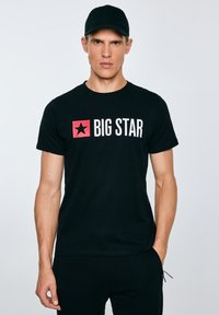 Férfi fekete sapkával és fekete pólóval, amelyen piros csillag és fehér "BIG STAR" felirat látható, egyik kezével a zsebében áll, egyszínű háttér előtt.