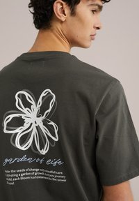 Olíva zöld t-shirt, nagy, fehér virág grafikával a hátsó részén, rajta a "élet kertje" felirattal. Pamut anyag, rövid ujjú.