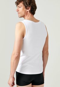 White cotton tank top with a classic round neck, fitted silhouette, and a smooth texture. Worn with black shorts, showing the back view.