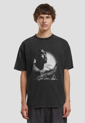 Młoda osoba z krótkimi kręconymi włosami, ubrana w czarny T-shirt z czarno-białym portretem i podpisem Tupaca Shakura.