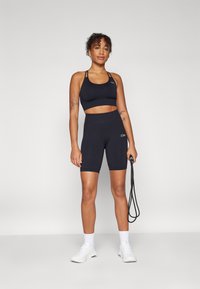 Crni atletski crop top i šortsevi visokog struka od rastezljivog materijala. Model drži konopac za preskakanje, nosi bijele tenisice s čarapama.