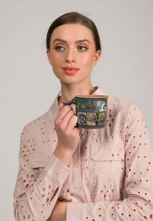 Młoda kobieta w jasnoróżowej bluzce z ażurowym wzorem, trzymająca kolorowy ceramiczny kubek z nadrukiem "Anekke".