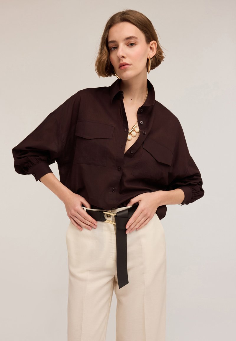 Maglietta marrone scuro dalla vestibilità morbida con due taschini sul petto, indossata con pantaloni color crema e una cintura nera con un accento dorato.