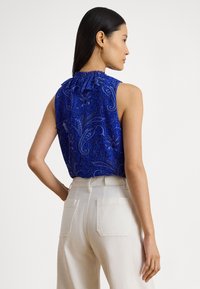 Donna con lunghi capelli scuri e orecchini a cerchio dorati che indossa un top senza maniche blu reale con motivo paisley e pantaloni bianchi a vita alta, di profilo.