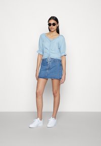 Top curto de manga curta, em azul claro com volantes e detalhe de cordão, combinado com uma saia mini de ganga. O modelo usa óculos de sol pretos e ténis brancos.