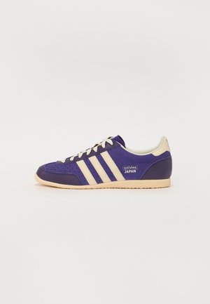 Niskoprofilowy sneaker Adidas Japan w ciemnym fioletowym kolorze z beżowymi paskami, sznurówkami, pętelką na pięcie i beżową gumową podeszwą, pokazany z boku.