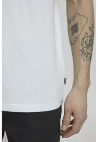 Hvid bomulds t-shirt med rund halsudskæring, korte ærmer, slidser i siden og et lille sort label i nederkanten; arm synlig med en tatovering.