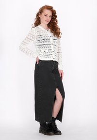 Weißer gestrickter Cardigan mit Lochmuster und Knöpfen, kombiniert mit einem schwarzen Denim-Maxikleid mit einem Oberschenkelspalt und schwarzen Stiefeln.
