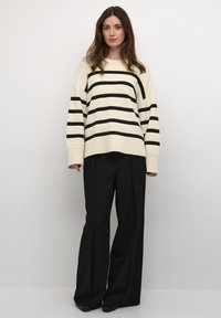 Übergroßer cremfarbener Pullover mit schwarzen Streifen und strukturiertem Strick, kombiniert mit weiten schwarzen Hosen und spitzen schwarzen Schuhen.