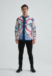 Homme debout portant une chemise à manches longues colorée et à motifs, un pantalon noir et des chaussures noires devant un fond léger uni.