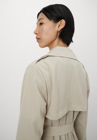 Beige trench coat featuring a double-breasted design, large collar, and belt. Smooth texture with a flowing silhouette and pleated back detail.