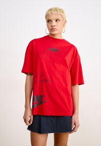 Raudona medvilninė marškinė su laisvu siluetu, su juodais Nike logotipo grafika ir tekstu priekyje. Strukturizuota medžiaga, trumpomis rankovėmis.