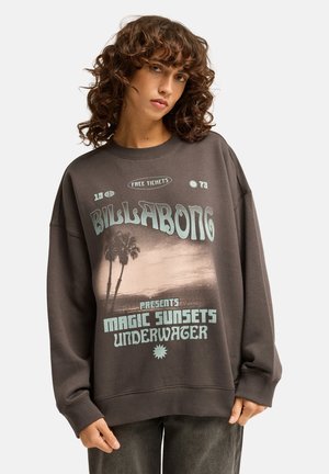 Kobieta z kręconymi włosami nosząca oversize ciemną bluzę z palmami i napisem Billabong Magic Sunsets Underwater.