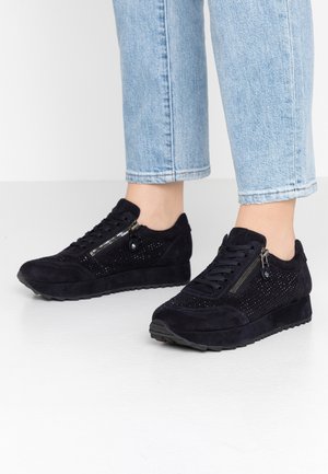 Person wearing black suede sneakers with side zippers and small perforations, paired with light blue cropped jeans.