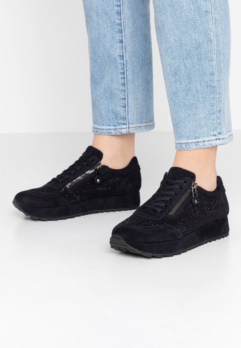 Personne portant des baskets en daim noir avec des fermetures éclair latérales et de petites perforations, associées à un jean court bleu clair.