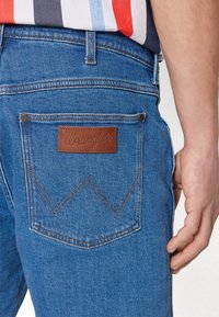 Modré džíny z denimu s koženou nášivkou "Wrangler" na zadní kapse, zdobené kontrastním hnědým šitím a charakteristickým zakřiveným designem.