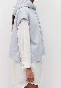 Světle modrá bezrukávová mikina z bavlněné směsi, se stahovací kapucí a žebrovaným lemem, přeložená přes bílou košili s dlouhým rukávem a knoflíkovými manžetami.