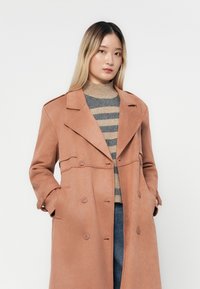 Cămașă trench roz pal cu un design cu guler dublu, guler decupat și buzunare frontale. Materialul pare moale, cu o textură netedă.