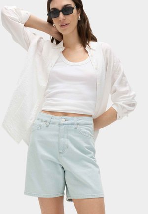 Mujer con gafas de sol negras, top blanco sin mangas, camisa blanca con textura y botones, pantalones cortos vaqueros azul celeste y pendientes de perlas.