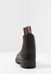 Bota de tornozelo em couro preto com painel lateral elástico amarelo, puxadores marrons, costura marrom e sola de borracha texturizada.