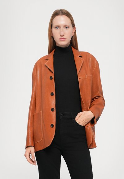 Brown leather blazer with a notched collar, black buttons, front pocket, and relaxed fit, worn over a black turtleneck and black pants.