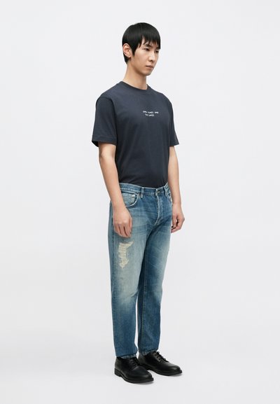 T-shirt bleu marine foncé avec texte blanc, jean clair bleu déchiré et chaussures noires à lacets. Le mannequin est debout, droit, sur un fond clair.