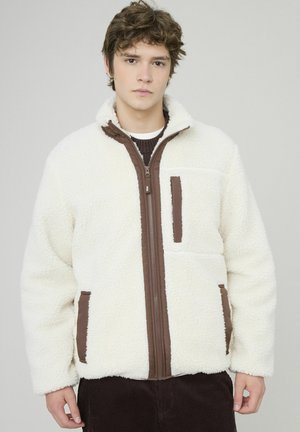 Veste en polaire blanche avec des accents de fermeture éclair marron à l'avant et sur les poches. Texture douce, coupe décontractée, col zippé et poches latérales.