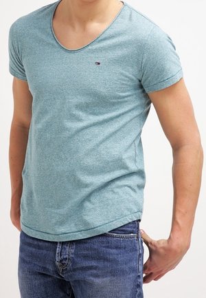 Mann trägt ein hellblaues kurzärmeliges V-Ausschnitt-T-Shirt und blaue Jeans, hat eine Hand lässig in der Nähe seiner Gesäßtasche ruhend.