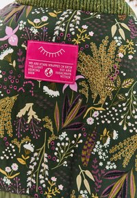 Zelená tkanina s různorodými květinovými motivy v růžové, bílé a zlaté barvě. Obsahuje jasně růžovou značku s bílým textem.