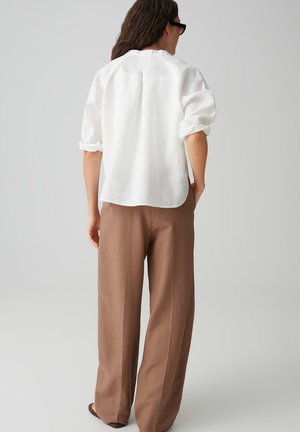 Persoon staand met de rug naar de camera, gekleed in een wit shirt met lange mouwen met opgerolde manchetten en losse bruine broek tegen een effen achtergrond.