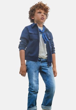 Giacca blu navy con zip e due tasche sul petto, t-shirt bianca, sciarpa blu e jeans con orlo arrotolato, in piedi su uno sfondo neutro.