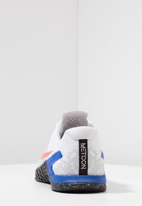 Zapatilla deportiva blanca con parte superior de malla texturizada, suela de goma negra, acentos azules y rojos, y una cinta en la parte trasera.
