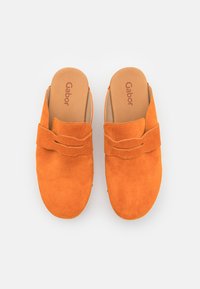 Des chaussures slip-on en daim orange avec une texture douce, présentant un large strap décoratif et une semelle intérieure rembourrée, conçues pour le confort.