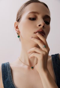 Gioielli in tono oro con gemme verdi a forma di cuore. La collana ha una catenina; gli orecchini e un anello completano il design.